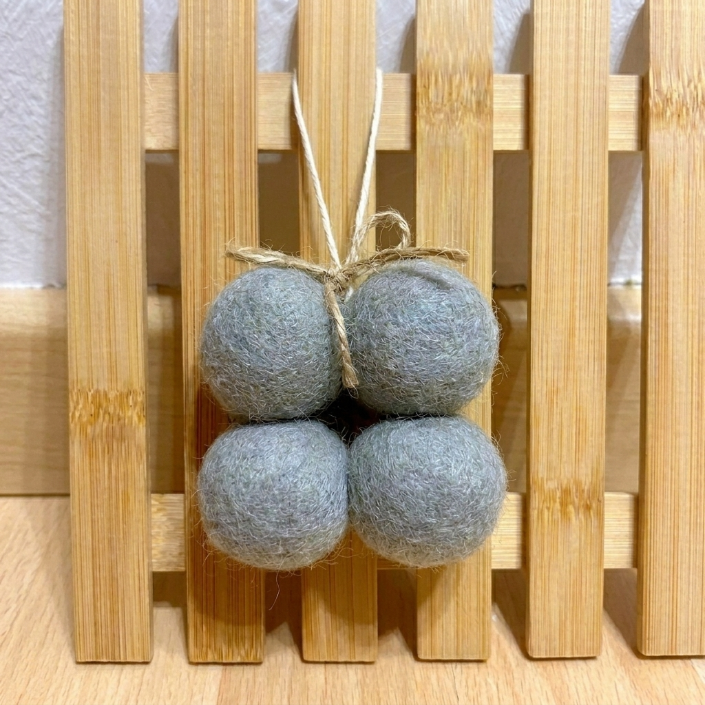 Minicolgante de cuatro bolas de fieltro de lana en color gris claro, colgado sobre un panel de bambú.